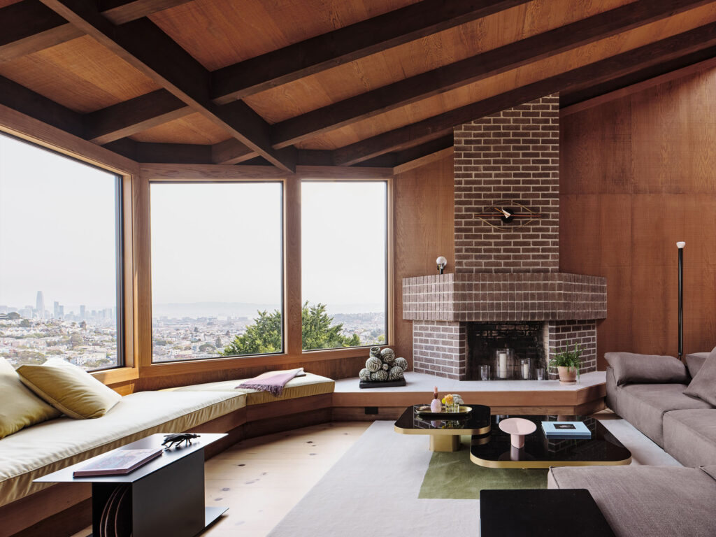 an image of a living room with large windows looking out to a city. There is a built in sofa along the windows that is connected to a brick fireplace