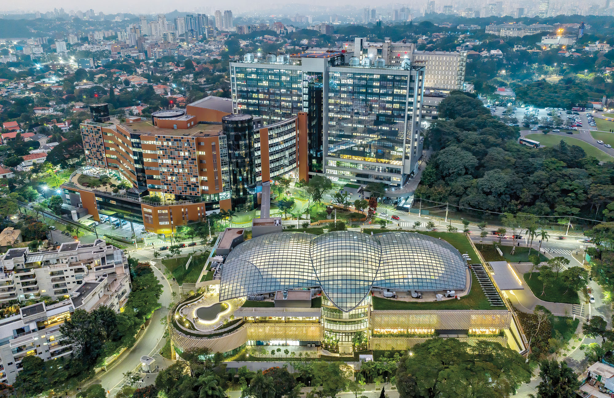 An aerial photograph of the Albert Einstein Education and Research Center designed by Moshe Safdie in Sao Paulo Brazil in the evening, it is illuminated from inside and surrounded by greenery.