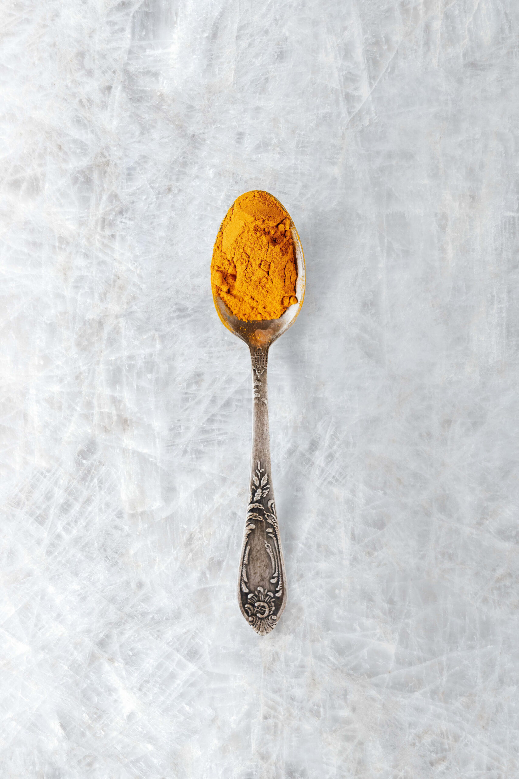 Silver spoon holding turmeric powder resting on white ceramic tile surface.