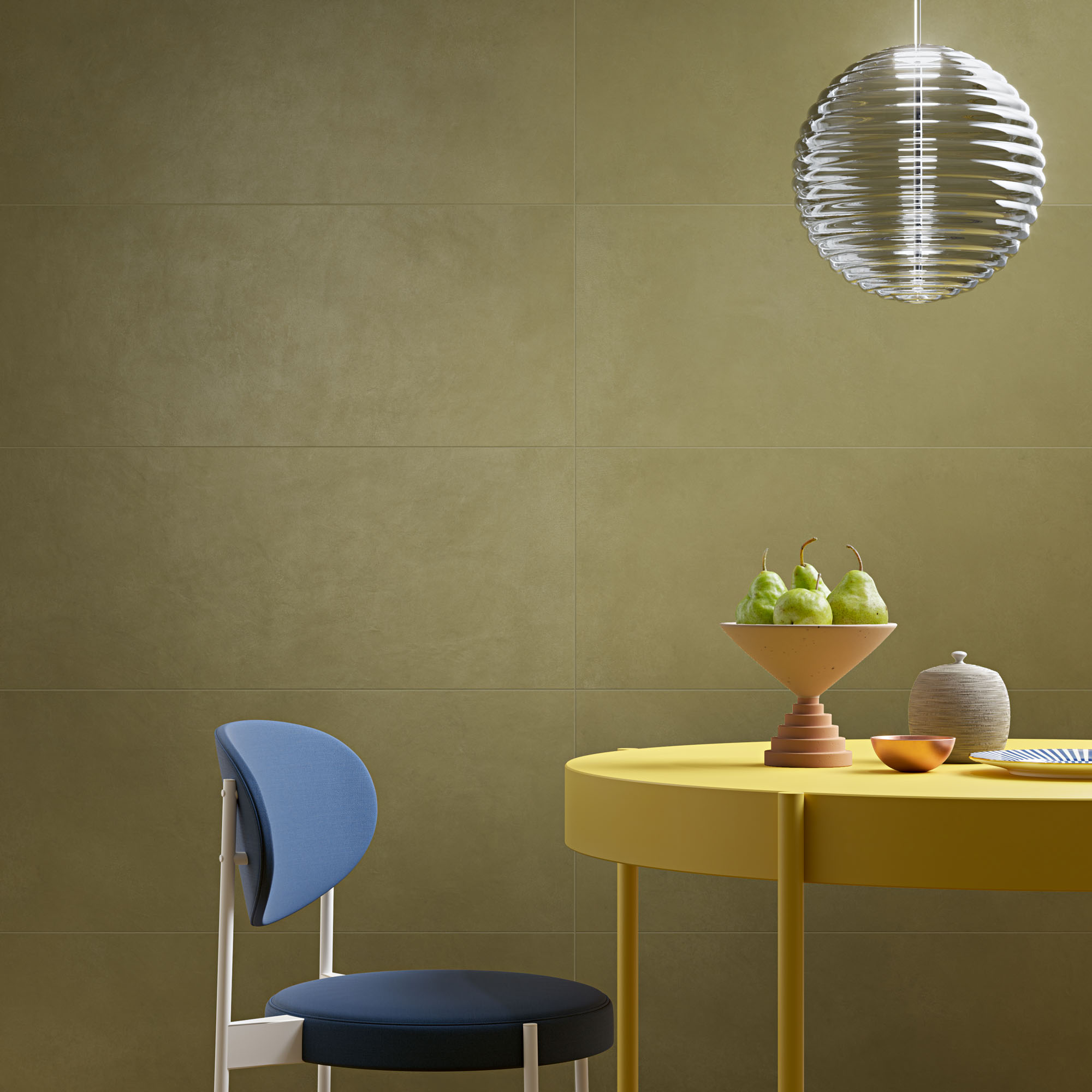Yellow table holding pears in a bowl and a blue chair with silver pendant light in front of mustard yellow ceramic tile-clad wall.