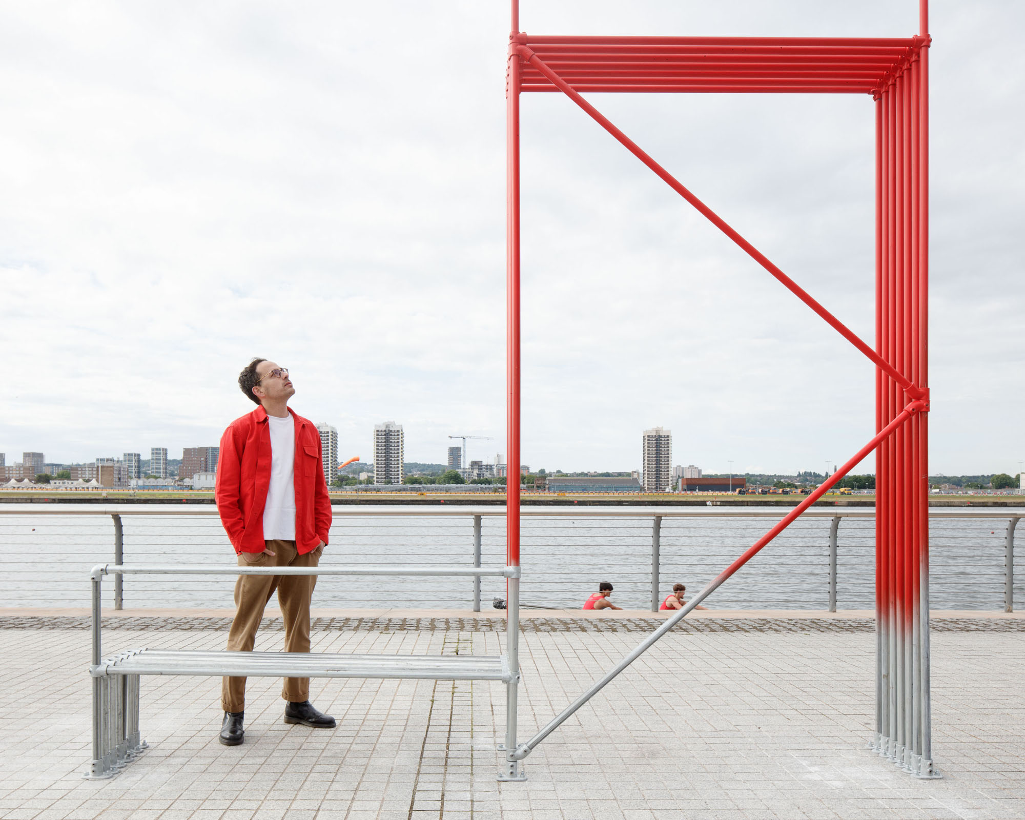 man in a red jacket standing next to a red sculpture by the water