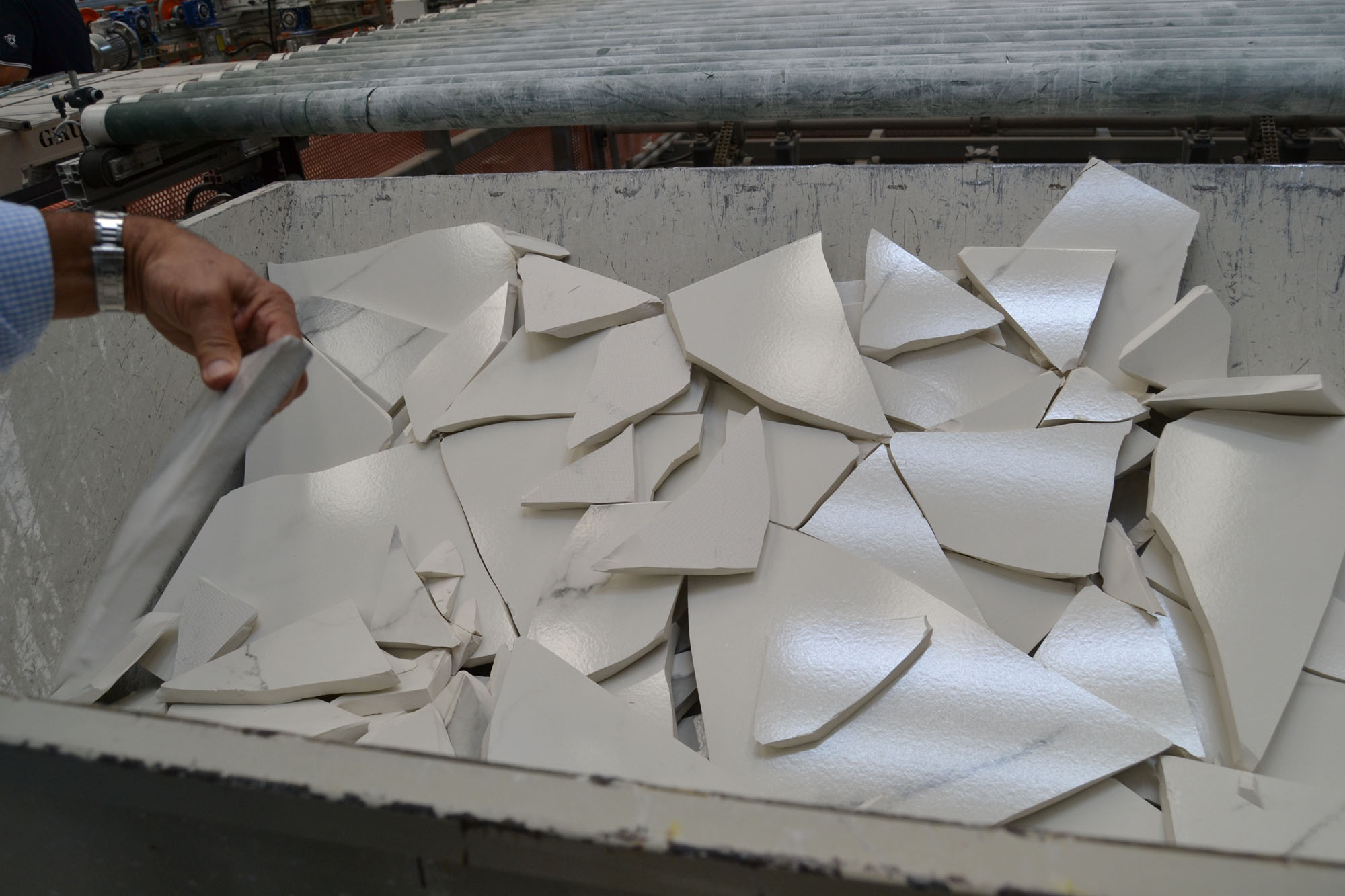 A hand picking up a shard of white, broken clay tile from a bin full of them.