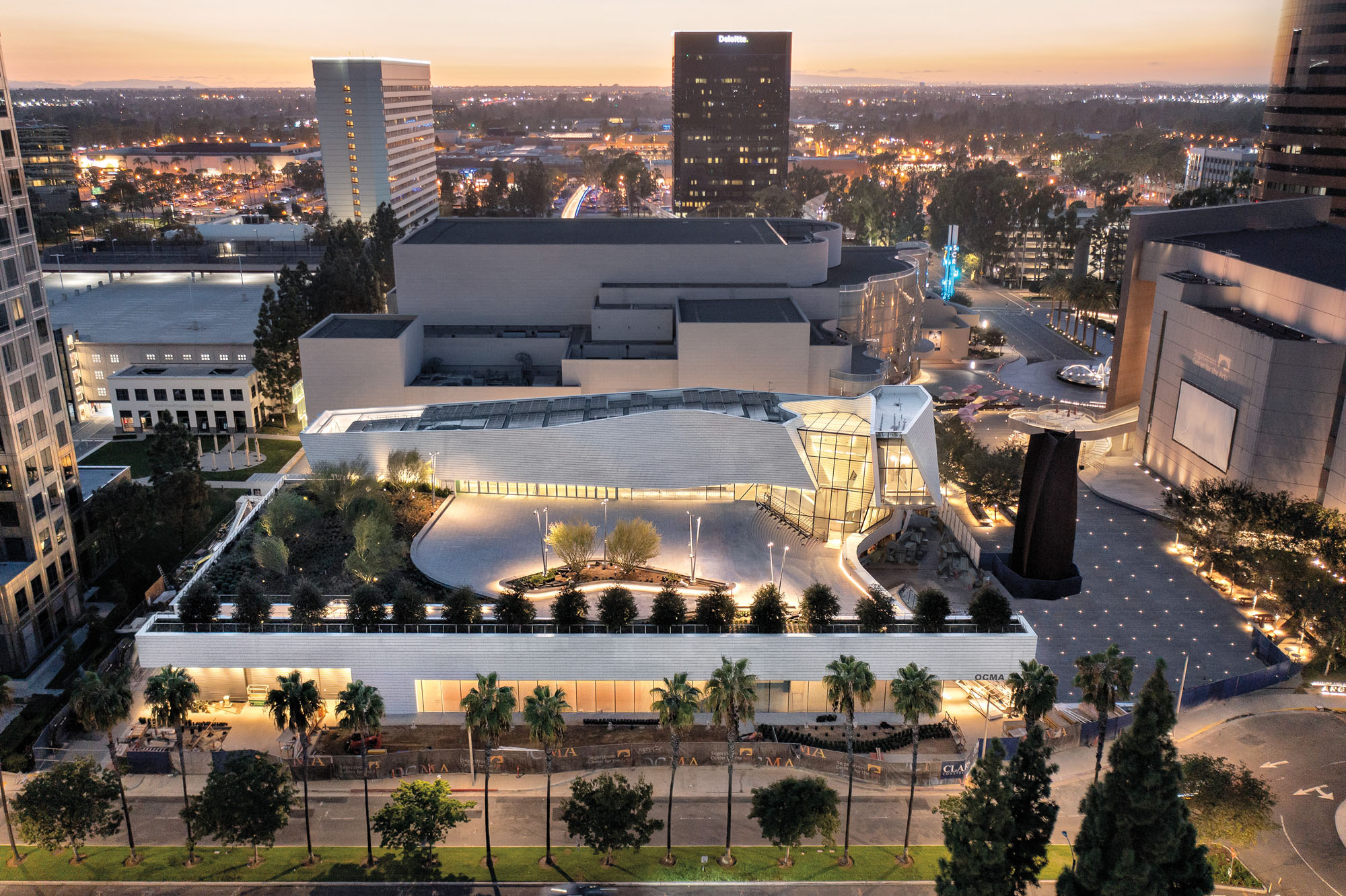 An aerial photograph of the Orange County Museum of Art in the evening shows roof garden and white facade.