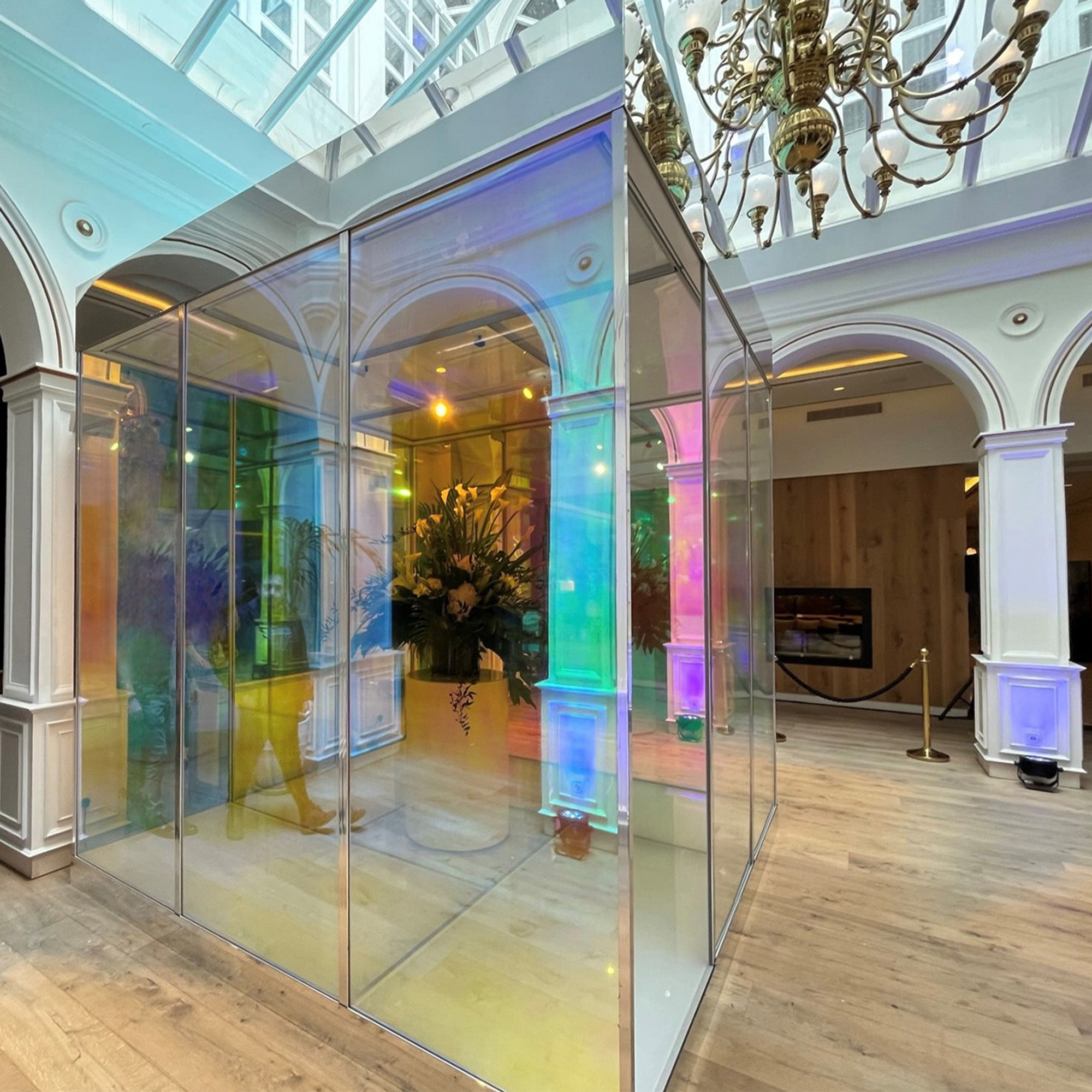 Clear glass room-size cube surrounded by round archways inside a building under a chandelier.