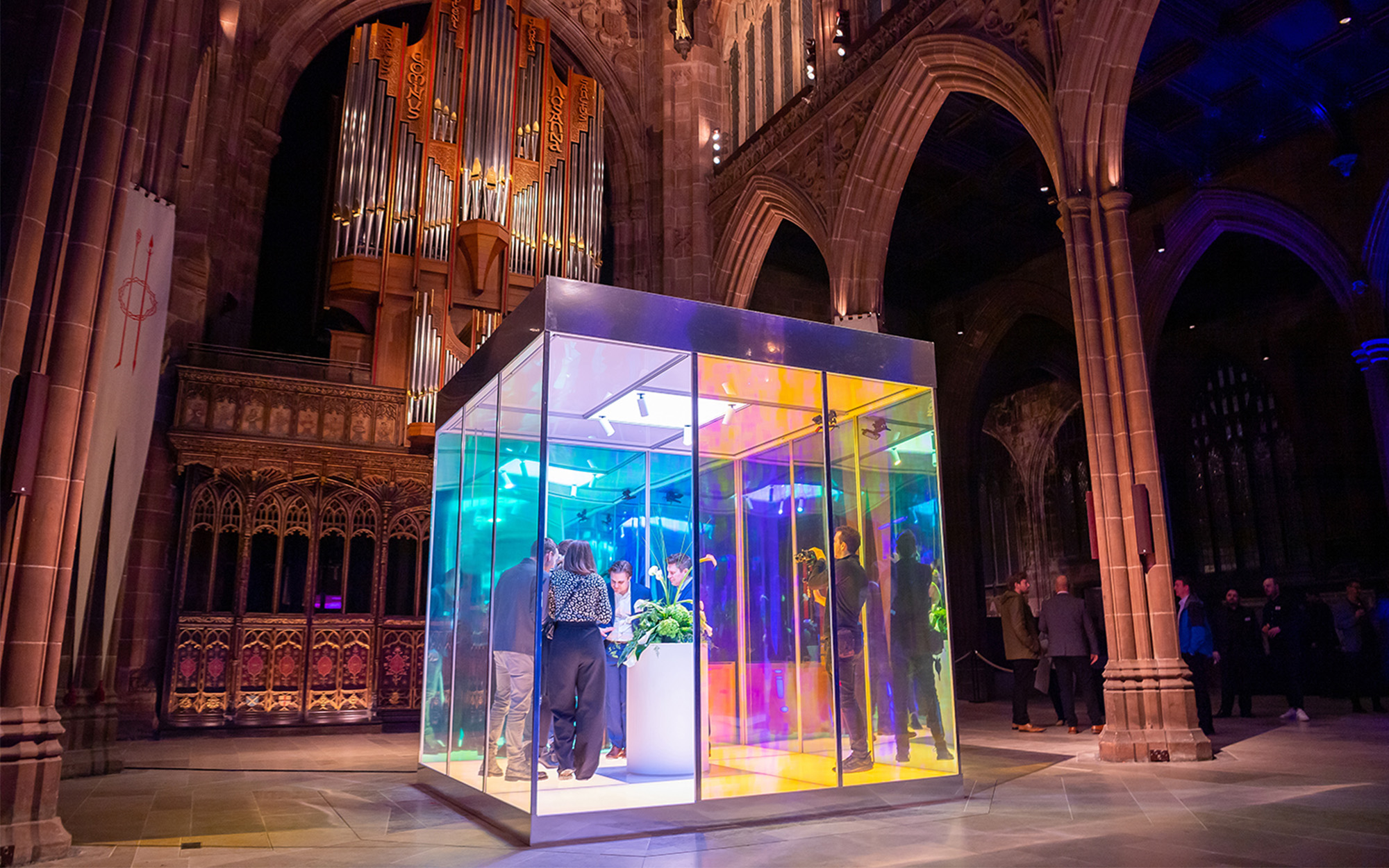 People inside of an illuminated glass cube, inside of a gothic cathedral with stone archways.