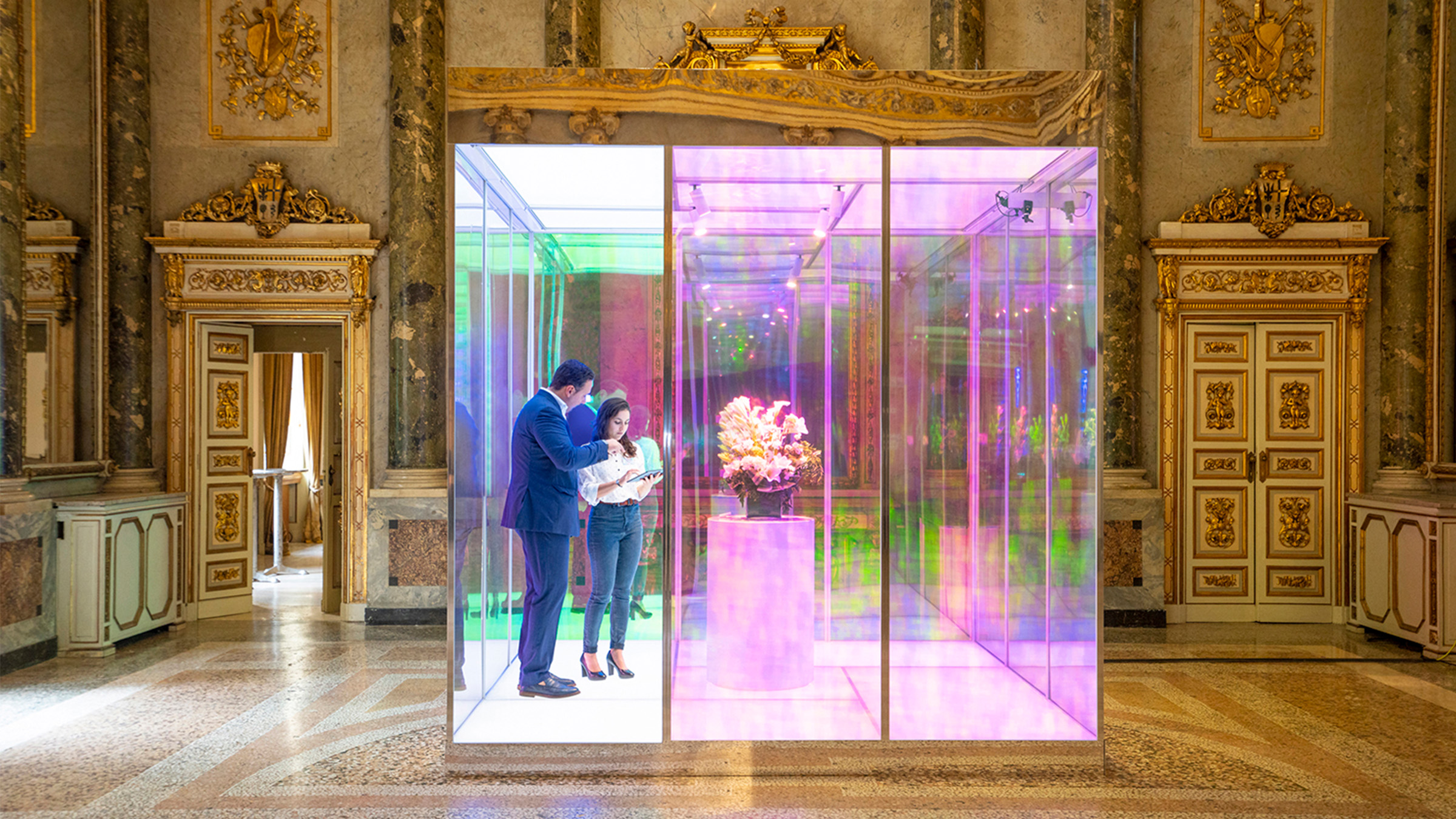 People in a glass cube in the middle of a baroque palace chamber surrounded by gilded, elaborately carved woodwork and terrazzo flooring.